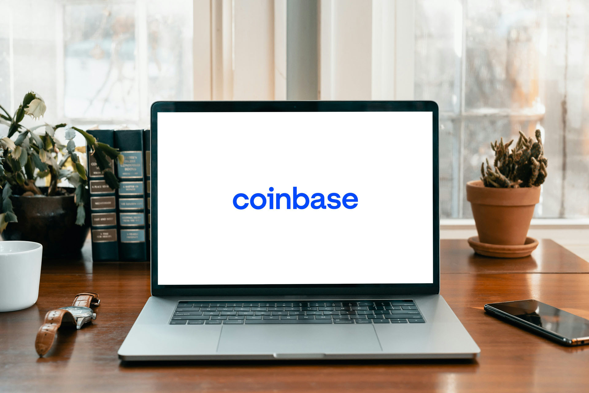 Coinbase logo displayed on a laptop screen in a cozy study setup with plants and books.