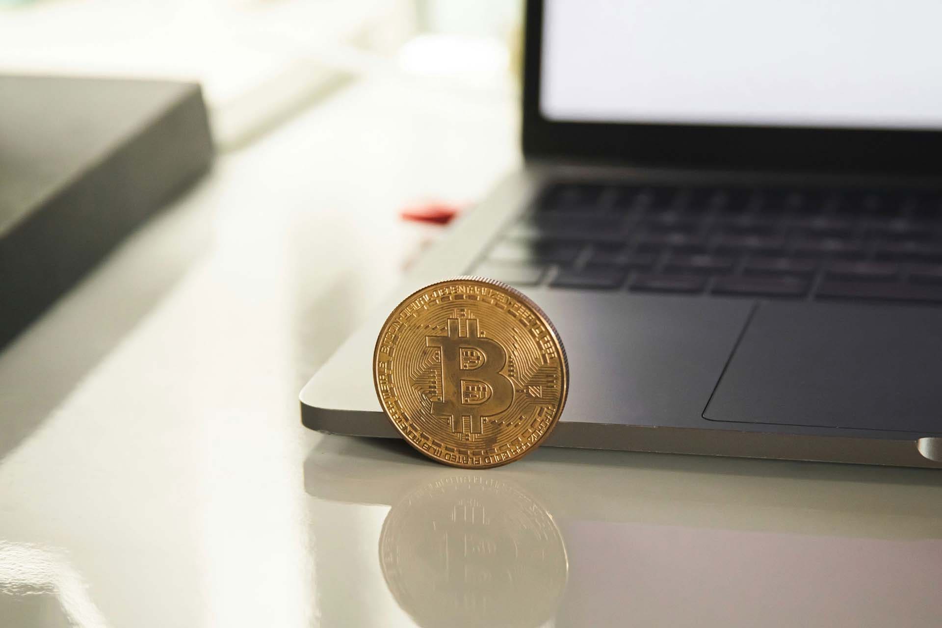 Bitcoin coin standing on the edge of a laptop, representing the intersection of digital currency and technology.